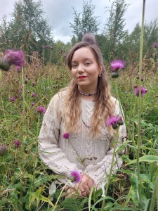Sunna Nousuniemi, a white-skinned person with long ash blonde hair sits in a meadow surrounded by brown knapweed flowers smiling and looking into the camera. Framed from the knees up, Nousuniemi is wearing a white cardigan, a colourful necklace and red lipstick.