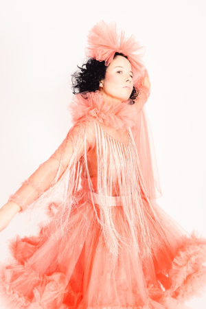 Jenni Laiti, a white-skinned person with black shoulder-length and curly hair, stands sideways in front of a white backdrop looking into the camera. Framed from the knees up, Laiti is wearing a salmon coloured gákti and a salmon coloured tulle headpiece.