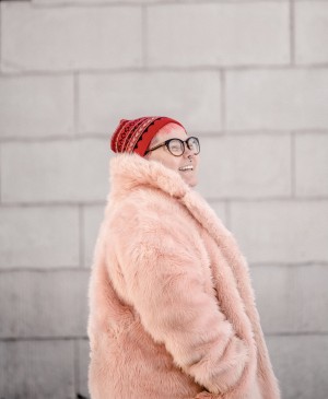 Merethe Kuhmunen, a white-skinned person with pink short hair stands in front of a gray brick wall smiling and looking to the right side of the camera. Framed from the waist up, Kuhmunen is wearing black and brown frame glasses, a red patterned headwear, and a light pink furry coat.