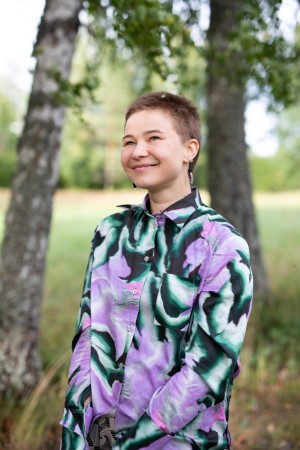 Vilma Leminen, a white-skinned person with short brown buzz cut, stands in the middle of two birch trees smiling and looking to the left corner of the camera. Framed from the waist up, they are wearing a colourful patterned blouse and earrings.