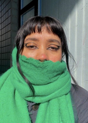 Amy R. Gelera, a brown-skinned person with black long hair, is standing in front of a white wall and door entrance looking into the camera. Framed from the chest up, Gelera is wearing a bright green scarf, black shirt and eyeliner.
